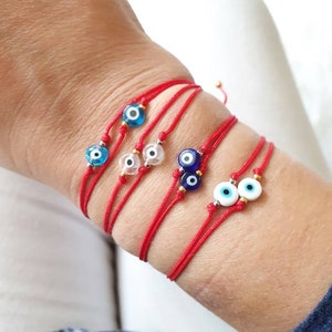 May include: Several red string bracelets with blue and white evil eye charms are displayed on a wrist. The bracelets have small gold-colored beads. The evil eye charms have a blue center with a black dot and white outer ring.