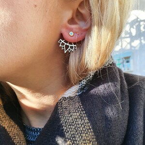 May include: A silver-coloured, lotus-shaped earring worn on the ear. The earring has a delicate, openwork design. The earlobe also has a small, round stud earring with a light blue centre. The person is wearing a dark-coloured scarf.