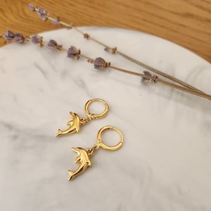 Könnte beinhalten: Ein Paar goldfarbene Creolen-Ohrringe mit Delfin-Motiv. Die Ohrringe sind auf einer weißen Marmoroberfläche mit getrockneten Lavendelzweigen im Hintergrund abgebildet. Die Ohrringe haben eine polierte Oberfläche.