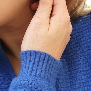 以下が含まれることがあります： 銀色のしずく型のイヤリング。イヤリングは滑らかで湾曲したデザインです。その人は青いセーターを着ています。イヤリングは、マニキュアを塗った手で触られています。