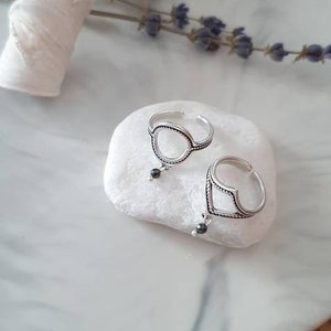 Puede incluir: Dos anillos ajustables de plata con cuentas negras colgantes descansan sobre una piedra blanca. Los anillos tienen diseños geométricos abiertos. La piedra está sobre un plato veteado, con lavanda seca y un elemento decorativo blanco.