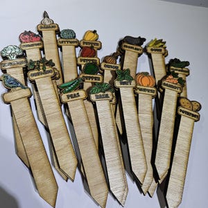 May include: Wooden garden markers with vegetable and herb illustrations. Each marker displays a plant name, such as "Garlic," "Lettuce," "Pepper," "Eggplant," "Corn," "Tomato," "Broccoli," "Oregano," "Peas," "Basil," "Mint," "Pumpkin," "Carrots," and "Potatoes."