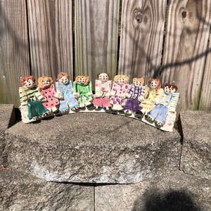 May include: A row of 12 ceramic figurines of people, each wearing different coloured clothing and sitting on a stone surface. The figurines are all facing forward and have painted faces.