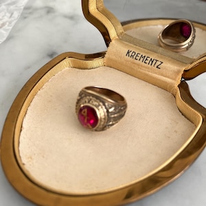 May include: A gold ring with a red gemstone sits in an open, gold-edged jewellery box. The ring features detailed patterns around the red stone. The box is lined with cream fabric, and the brand name "Krementz" is printed on the inside of the box.