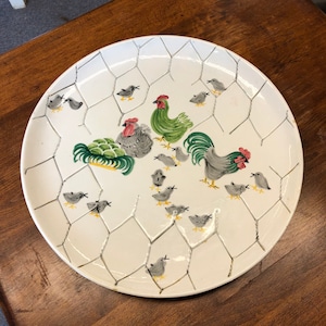 May include: A white ceramic serving platter with a chicken coop design. The platter features a rooster, hens, and chicks, all surrounded by a wire fence pattern. The chickens are painted in shades of grey, green, and red.
