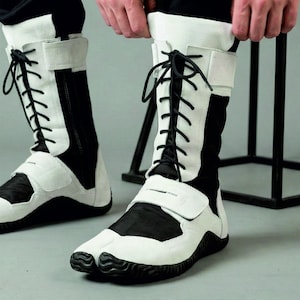 May include: Black and white split-toe boots with lace-up closures and Velcro straps. The boots have a high-top design with a white upper and black accents. The soles are black and textured.
