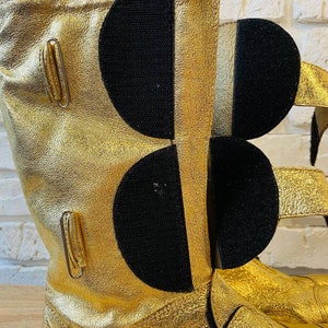 May include: Gold leather boots with black velcro straps and buckles. The boots have a textured surface and a pointed toe.