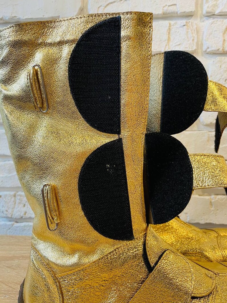 May include: Gold leather boots with black velcro straps and buckles. The boots have a textured surface and a pointed toe.