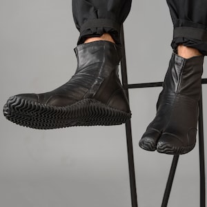 May include: Black leather ankle boots with a split toe design and a thick rubber sole. The boots are worn by a person sitting on a chair.