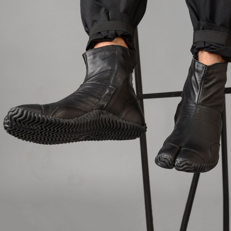 May include: Black leather ankle boots with a split toe design and a thick rubber sole. The boots are worn by a person sitting on a chair.
