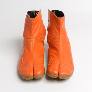 Op de afbeelding: Een paar oranje leren tabi-laarzen met een gespleten teen. De laarzen hebben een textuurzool en een rits aan de achterkant.