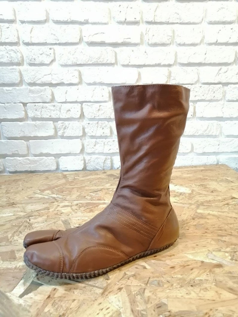 Puede incluir: Botas de cuero marr&oacute;n con un dise&ntilde;o de punta dividida. Las botas tienen un eje alto y una punta redonda.