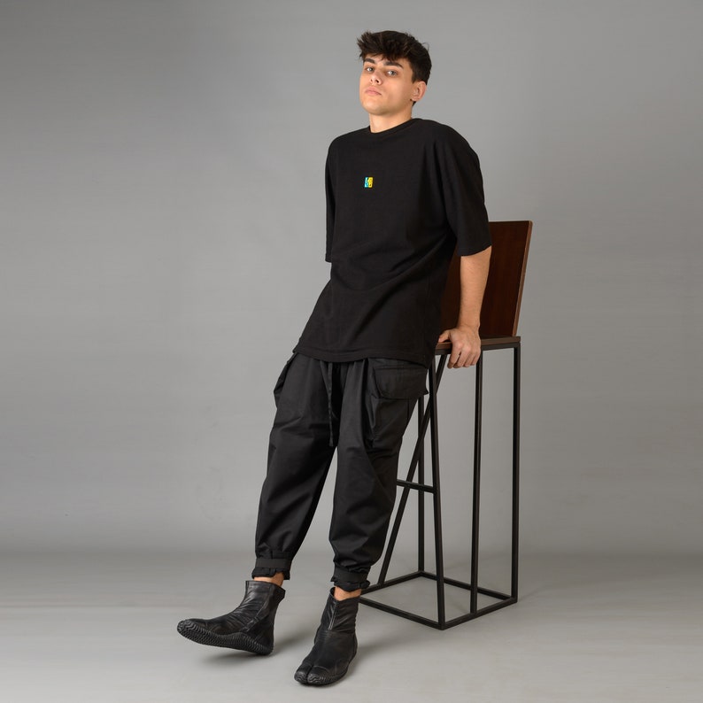 May include: A person wearing a black t-shirt with a small colorful design, black pants, and black shoes. The person is leaning against a black metal stool with a wooden top.