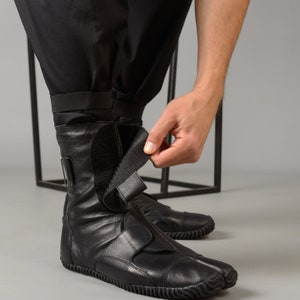 May include: Black leather ankle boots with a unique toe design and adjustable straps. The boots have a rubber sole and are being worn by a person.