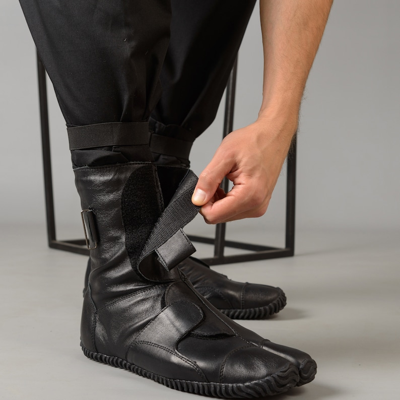 May include: Black leather ankle boots with a unique toe design and adjustable straps. The boots have a rubber sole and are being worn by a person.