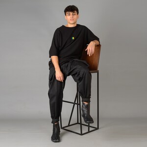 May include: A person wearing a black t-shirt with a small colorful design sits on a black metal stool with a wooden seat.