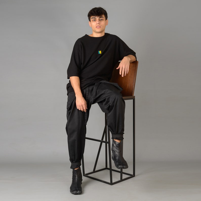 May include: A person wearing a black t-shirt with a small colorful design sits on a black metal stool with a wooden seat.