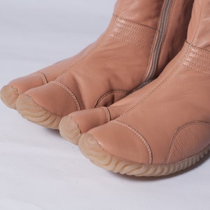 Puede incluir: Un par de botas tabi de cuero rosa claro con una cremallera en la parte posterior. Las botas tienen una suela de goma y un dise&ntilde;o de punta dividida.