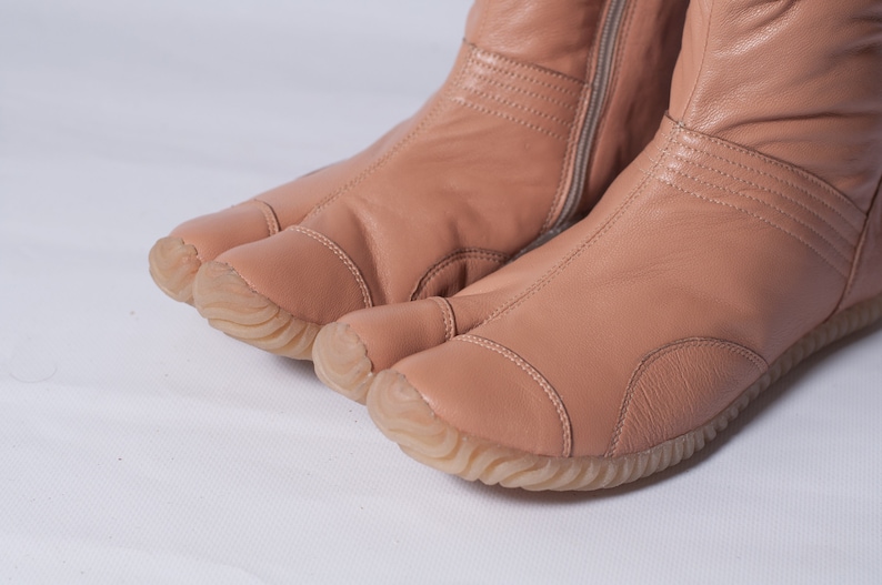 Puede incluir: Un par de botas tabi de cuero rosa claro con una cremallera en la parte posterior. Las botas tienen una suela de goma y un dise&ntilde;o de punta dividida.