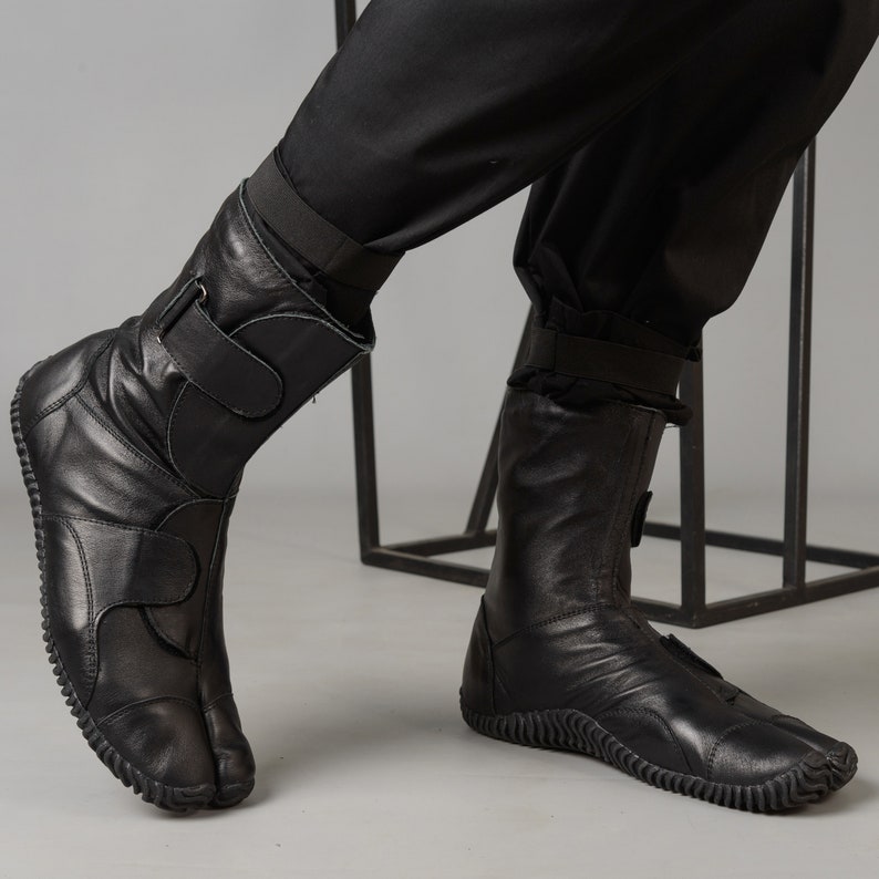 May include: Black leather boots with a split toe design and a strap across the ankle. The boots have a thick rubber sole.