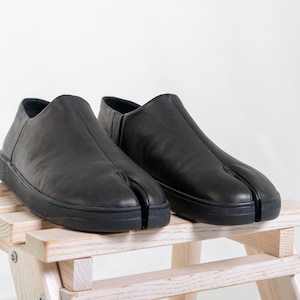 Op de afbeelding: Een paar zwarte leren schoenen met split-teen en een laag profiel. De schoenen worden gepresenteerd op een lichtgekleurde houten kruk tegen een witte achtergrond. De schoenen hebben een uniek ontwerp met een apart gedeelte voor de grote teen.