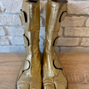 May include: A pair of gold leather boots with a split toe design. The boots have a buckle closure and a textured sole.