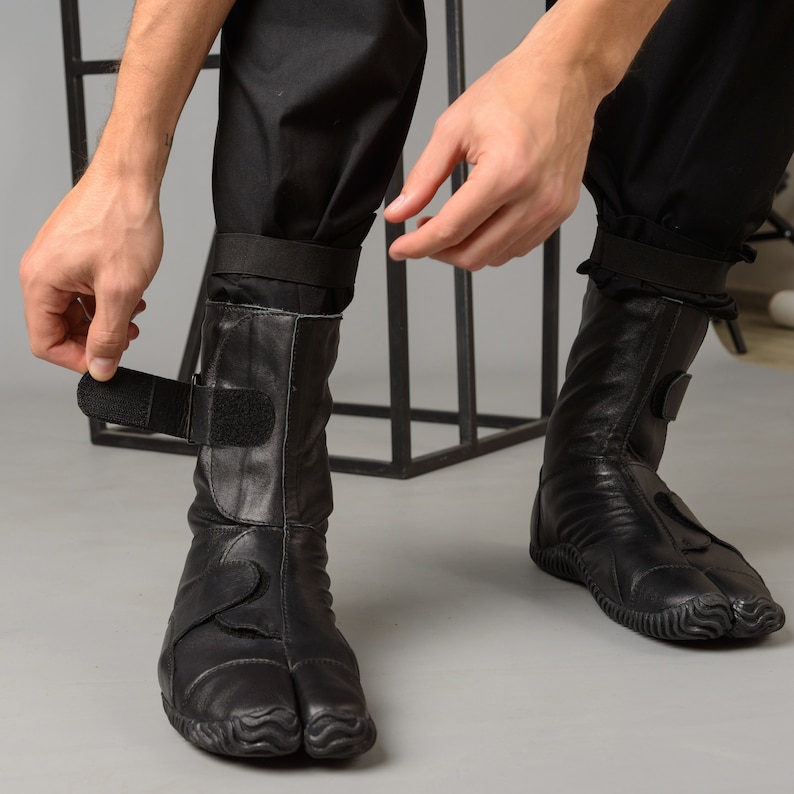 May include: A pair of black leather boots with a split toe design and adjustable straps. The boots have a textured sole and are worn on a person's feet.