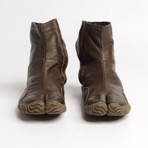 May include: A pair of brown leather tabi boots with a split toe design. The boots have a textured rubber sole and a zipper on the back.