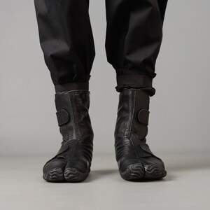 May include: A pair of black leather boots with a unique, textured sole. The boots have a strap across the ankle and a zipper on the side.