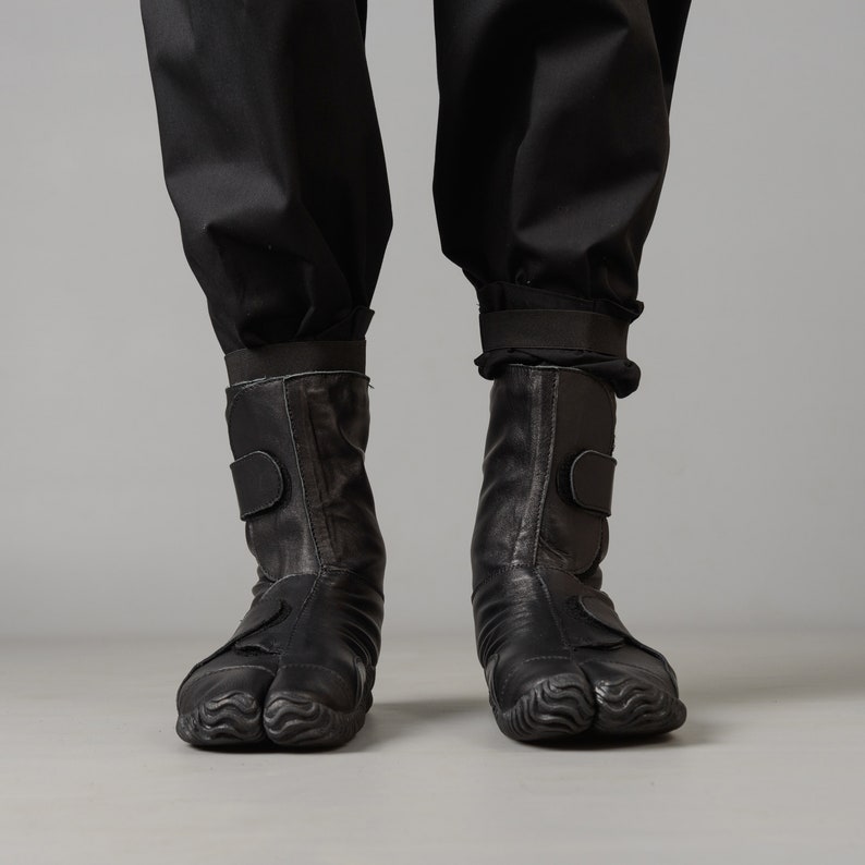 May include: A pair of black leather boots with a unique, textured sole. The boots have a strap across the ankle and a zipper on the side.