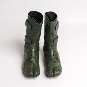 Puede incluir: Un par de botas de cuero verde oscuro con m&uacute;ltiples correas y un dise&ntilde;o de punta dividida.