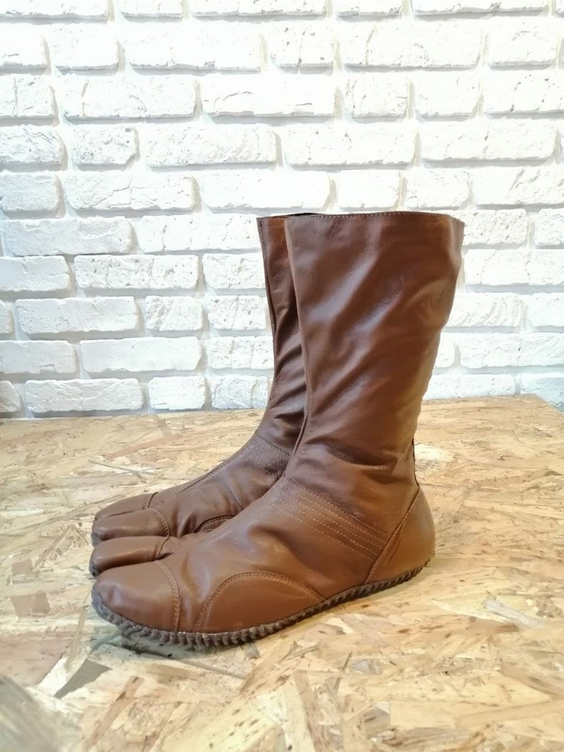 Puede incluir: Un par de botas de cuero marr&oacute;n con un dise&ntilde;o de punta dividida. Las botas tienen un estilo simple y minimalista y est&aacute;n hechas de un cuero suave y flexible.