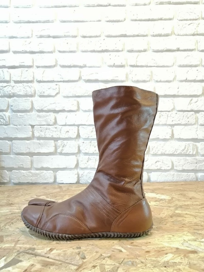 Puede incluir: Botas de cuero marr&oacute;n con un dise&ntilde;o de punta dividida. Las botas son de altura por encima de la rodilla y tienen un estilo simple y elegante.