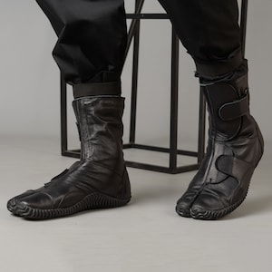 May include: Black leather boots with a unique split-toe design and a strap at the ankle. The boots have a textured rubber sole.