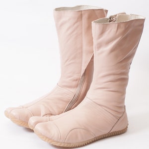 Puede incluir: Un par de botas de cuero rosa claro con una cremallera lateral y un dise&ntilde;o &uacute;nico de punta dividida.