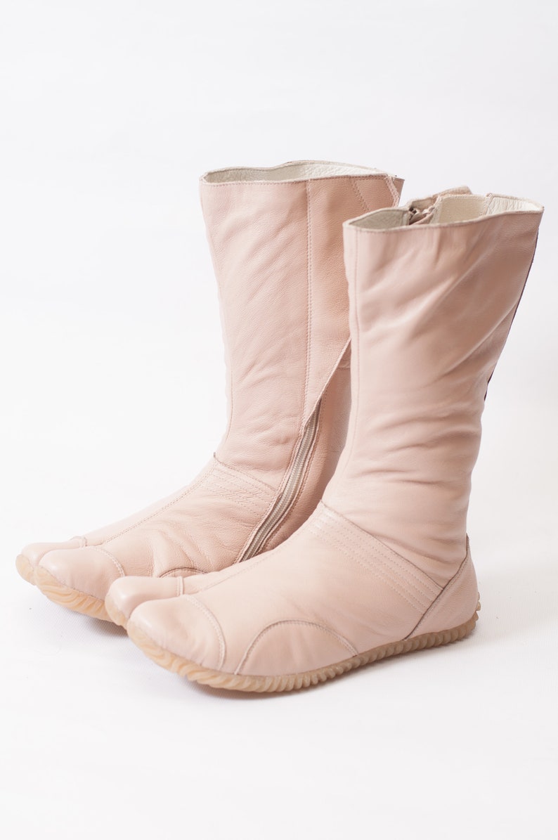 Puede incluir: Un par de botas de cuero rosa claro con una cremallera lateral y un dise&ntilde;o &uacute;nico de punta dividida.