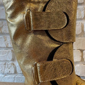 May include: Gold leather boots with two buckles and a decorative cut-out design.