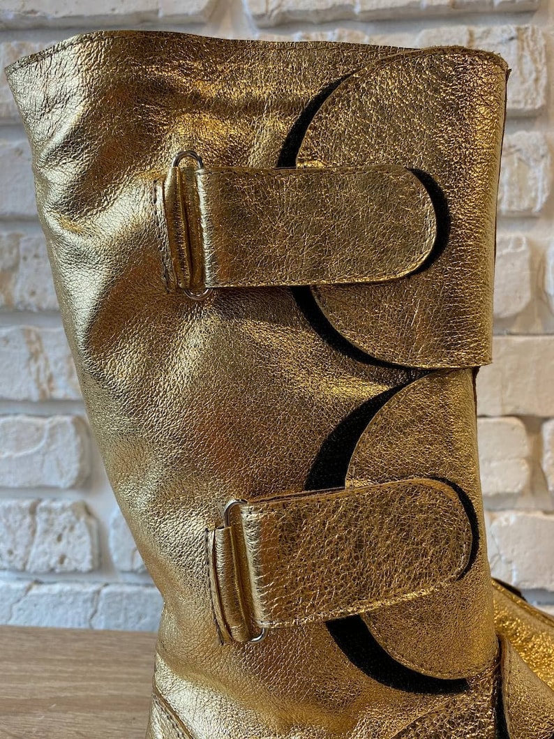 May include: Gold leather boots with two buckles and a decorative cut-out design.