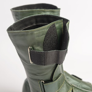 Puede incluir: Un par de botas de cuero verde con correas y hebillas negras. Las botas tienen una cremallera en el lateral y una punta puntiaguda.