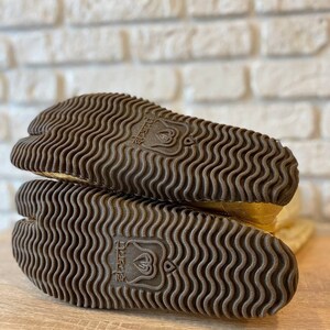 May include: Close-up of the bottom of a pair of gold tabi boots with a brown rubber sole. The sole has a wavy pattern and the text "tabi" is printed on the sole.