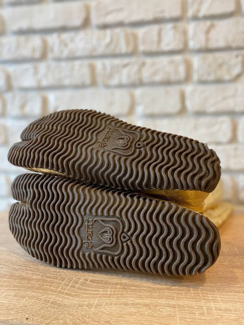 May include: Close-up of the bottom of a pair of gold tabi boots with a brown rubber sole. The sole has a wavy pattern and the text "tabi" is printed on the sole.