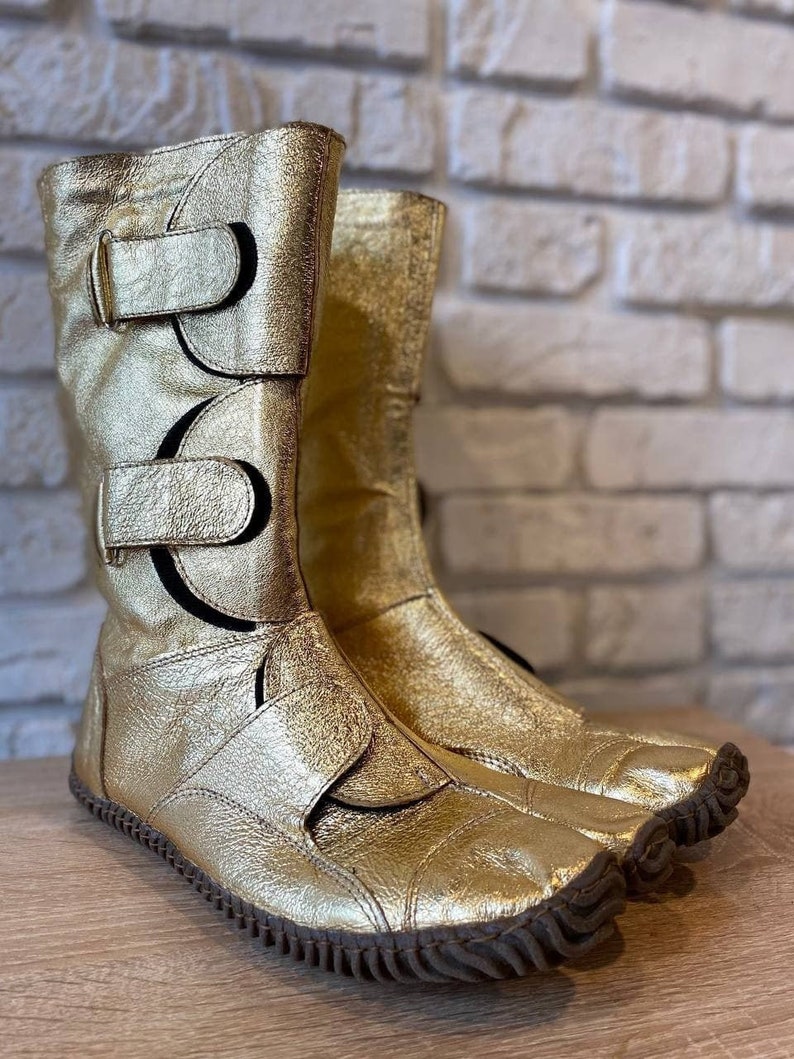 May include: A pair of gold leather boots with black buckles and a unique toe design. The boots have a textured sole and are made of a soft, supple leather.