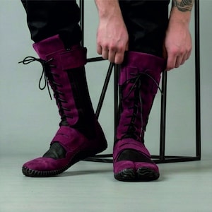 Op de afbeelding: Een paar hoge laarzen met split-teen in een diepe paarse tint. De laarzen hebben een vetersluiting met zwarte veters, een gespsluiting en een zwart paneel aan de voorkant. De zolen zijn zwart en getextureerd.