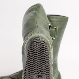 Puede incluir: Un par de botas de cuero verde con suela de goma negra. Las botas tienen cierre de hebilla y punta puntiaguda.