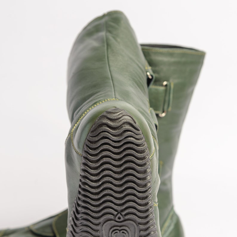 Puede incluir: Un par de botas de cuero verde con suela de goma negra. Las botas tienen cierre de hebilla y punta puntiaguda.