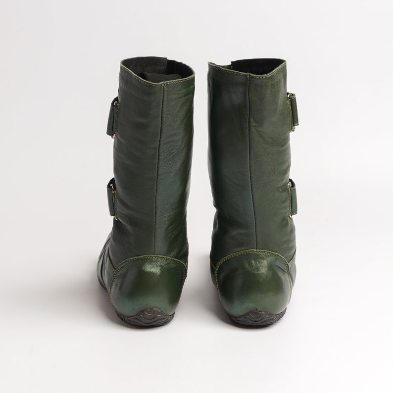 Puede incluir: Un par de botas de cuero verde oscuro con hebillas. Las botas tienen una punta redonda y un tac&oacute;n bajo.