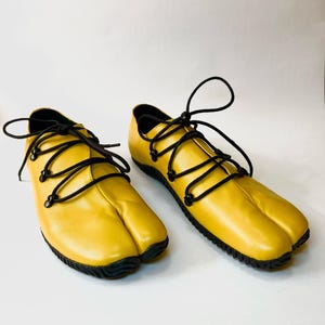 May include: A pair of yellow leather split-toe shoes with black laces. The shoes have a rubber sole and a rounded toe.