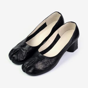 May include: A pair of black leather heeled shoes with a split-toe design. The shoes have a low block heel and a rounded toe. The interior is white. The shoes are displayed on a white background.