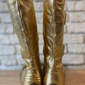 May include: A pair of gold leather boots with a buckle detail on the side. The boots have a textured rubber sole.