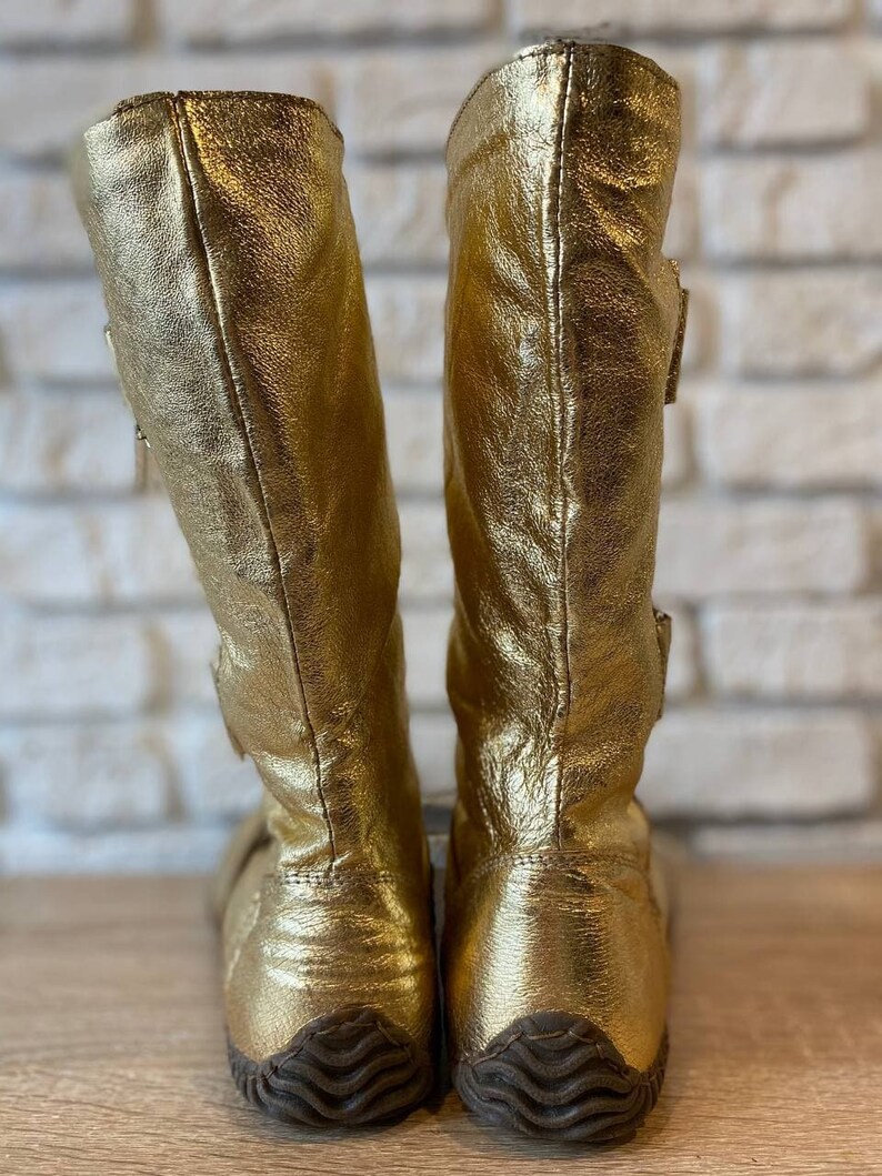 May include: A pair of gold leather boots with a buckle detail on the side. The boots have a textured rubber sole.